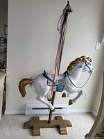 Side view of the white wooden carousel horse with gold mane and tail on a wooden cross base standing indoors.