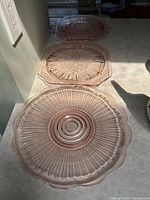 Three pink depression glass plates lined up on a counter, showing circular ribbed and floral pressed patterns with scalloped edges.