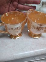 Front view of two peach-colored Fire-King cups with leaf embossing and footed bases, held by hand for scale.