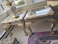 Photo showing two matching glass-top side tables with cream-colored carved wooden legs and detailed trim.