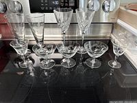 Photo showing nine assorted glassware items arranged in two rows: two crystal wine glasses, two champagne flutes, and five cocktail/dessert glasses with etched designs.