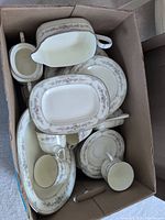 Box containing multiple pieces of bone china including cups, saucers, gravy boat, and serving dishes with floral pattern and silver trim