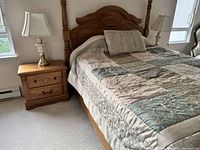 Queen bed with patchwork quilt, decorative pillow, and two wooden side tables each with brass and glass lamps and two drawers. Carpeted room with windows in background.