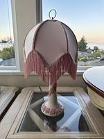 Full view of pink fabric lamp shade with fringe and ceramic base with grapevine decoration.