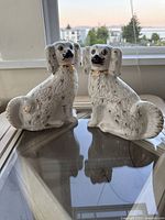 Two white porcelain dog statues with gold painted embellishments sitting on a glass table in front of a window.