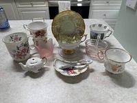 Photo of assorted china cups, saucers, a small teapot, pink glass dish, and plate on table