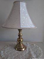 Photo of one brass base lamp with off-white floral fabric lampshade on white crocheted cloth