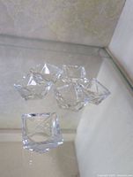 Top view of six small clear crystal square candle holders arranged in a flower shape on a glass surface showing their faceted sides and clarity.