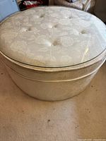 Full view of round coffee table with ivory floral upholstery and clear glass top, showing button tufting and some staining on the fabric.