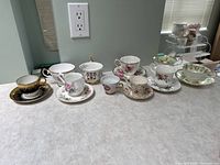 Photo showing all seven vintage bone china teacups with matching saucers arranged on a countertop, highlighting varied floral and gold-accented designs.