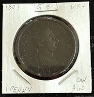 Obverse side of the 1807 Great Britain King George III one penny copper coin mounted in a white cardboard holder with the inscription '1807 G.B. VF 1 PENNY CAN $60' written on the holder.
