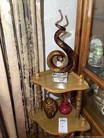 Three pieces of art glass displayed on a small wooden corner shelf, featuring an amber spiral sculpture on a clear base, a smaller amber swirled glass piece, and a small red vase-shaped glass piece.