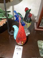 Red-bodied art glass rooster with blue comb and tail visible, positioned beside the speckled rooster.