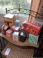Photo showing several vintage Avon collectible boxes arranged on wicker shelf by window. Different box designs, including 'Super Shoe' basketball box and others with floral and geometric patterns.