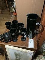 Photo showing set of 6 black amethyst glass goblets, one tall pitcher with clear handle, and a black glass leaf-shaped dish on wooden surface.