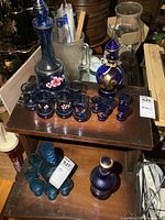 Photo showing four blue glass decanters and matching glasses on two wooden trays, along with other objects in the background.