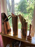 Full view of all brass and copper items on shelf with window background, showing the pitcher, match holders, and bud vase.