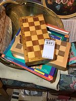 Three wooden chess boards stacked with one partially inside a wooden box, placed on top of chess and game-related books and materials.