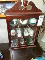 Front view of wooden collectibles cabinet showing painted eggs, porcelain pieces, and art glass figurines on shelves.