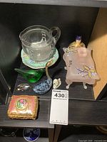 Photo showing various small collectibles arranged on black shelving, including glass pitcher, iridescent plate, bird figurines, small dog, metal dolphin, pink footed trinket box, and ornate metal trinket box.