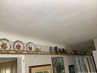View of 22 collector plates displayed on a high shelf mostly showing floral and scenic designs in a row.