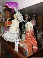 Overview of six dolls, including boxed plastic doll and porcelain dolls in vintage attire, displayed on top of a dark wood cabinet.