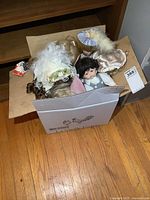 Photo of a cardboard box filled with multiple porcelain dolls, some with white hair and hats, showing the variety and condition of the items.