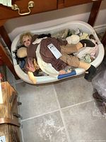 Basket full of dolls including a large cloth doll on top and several porcelain dolls beneath in detailed clothing.