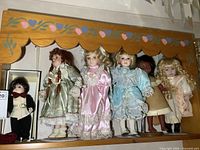 Photo showing six porcelain dolls arranged on a wooden shelf with painted floral decoration on the shelf edge, five female dolls in pastel dresses and one male doll in suit.
