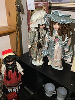Three porcelain dolls on a dark surface, one dressed in a blue satin dress, one in a checked dress with lace bonnet, and another in red folkloric attire with a yellow hat.