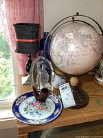 Globe, candle lamp, plate, blue pottery vase, Jim Beam bottle, and glass graduation figurine on wooden surface near window with pink curtains visible