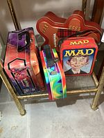 Four lunch boxes and tins including MAD magazine, Pokemon, a holographic lunch box, and a Walkers biscuit tin shaped like a dog bone.