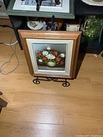 Framed signed oil painting of white and orange flowers in a vase on a metal stand on wooden floor.
