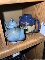 Two full-size pottery teapots on a wood shelf, one blue and cream with butterfly decoration, the other beige with brown accents.