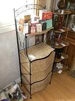 Front view of metal frame shelf with three woven rush drawers and items on top shelf, placed near window and wooden furniture.