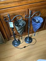 Pair of metal table lamps standing side by side on a wood floor with a carved wood cabinet background. One lamp has a gray spiral shade, the other a blue spiral shade. Both lamps have visible bulbs and cords with inline switches.