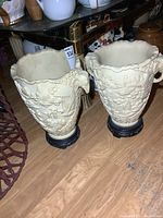 Pair of cream-colored resin urns with textured relief designs, elephant trunk handles, on wooden floor, one handle damaged