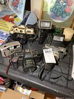Four vintage film cameras and two flash units arranged on a table. Cameras appear dusty and used. One camera is gold colored, one is a Vivitar compact model, two are black with larger lenses. Two flash units, one handheld with grip, one smaller, are included.