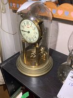 Vintage Kundo brass anniversary clock under glass dome, showing clock face and brass components on a black surface with a wooden heart-patterned background.