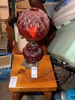 Full view of vintage red glass electric lamp on wooden table with ornate metal base, red glass globe top and bottom sections.
