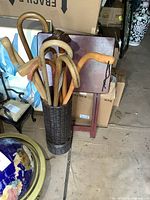 Wicker cane stand filled with several wooden canes in different styles and finishes, situated on a concrete floor near other furniture items.