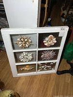 Front view of white wooden window frame with six glass panes containing shell art in floral/starburst patterns.
