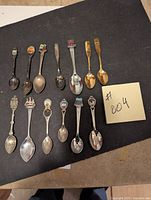 All 13 collector spoons arranged on black background showing different colors and designs including gold and silver tones and colored enamel emblems