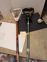 Two lacrosse sticks side by side on a table, one with a white head and orange shaft and the other with a black head and black shaft with yellow markings.