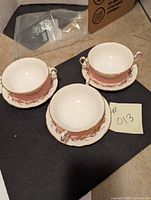 Three cream soup bowls with matching saucers shown from the top and side angles. Bowls have pink and gold leaf pattern near rim and single loop handle.