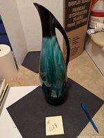 Blue Mountain Pottery jug viewed from an angle showcasing its tall slender shape, turquoise and dark brown gradient glaze, and handle.