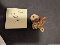 Photo of a brass Coca-Cola bottle opener with embossed brand name viewed from the front showing mounting holes and opener lip.