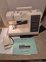 Front view of a white Kenmore sewing machine showing the stitch selection panel, foot pedal, and owner's manual beside it.