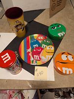 Photo showing five M&M branded items: one round plate with multi-colored characters, two round tins (green and orange) with character faces on lids, one red square tin with a coin slot on lid, and a tall yellow cup with an M&M character graphic.