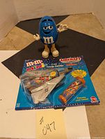 Packaged M&M Minis jet-shaped candy carrier with matching candy tube, displayed with blue M&M dispenser in background.